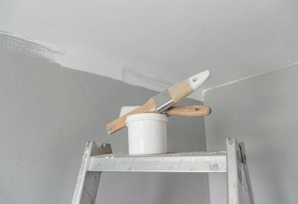A sash brush resting on a paint pot on a ladder shelf below a crisp ceiling cut line.