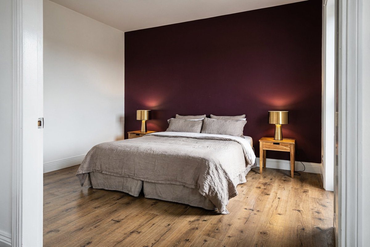 Master bedroom with deep burgundy feature wall, king bed with taupe linen bedding, brass lamps and oak floors in a renovated Williamstown home